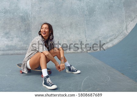 Portrait Of Skater Girl In Skatepark. Female Teenager In Casual Outfit Sitting On Skateboard Against Concrete Wall. Summer Skateboarding With Modern Sport Equipment As Part Of Active Lifestyle.
