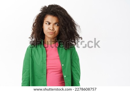 Portrait of skeptical young girl, student looks disappointed and gloomy, smirks as looks unimpressed, white background.