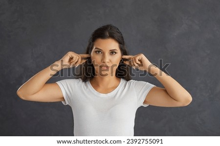 Portrait of skeptical young woman covering her ears with her fingers so as not to hear anything. Close up portrait of woman who is looking at camera with serious expression. Concept of human emotions.