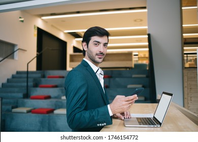 Portrait of skilled corporate employee looking at camera during work time for using modern technology, Indian male entrepreneur with digital smartphone gadget standing bear desk with mockup netbook