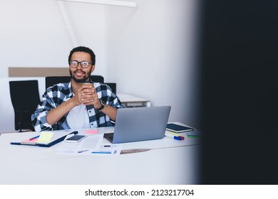 Portrait Of Skilled Employer In Eyeglasses Enjoying Coffee Break In Office Interior With Laptop Computer, Successful Spanish Businessman With Takeaway Cup Posing At Desktop With Digital Netbook