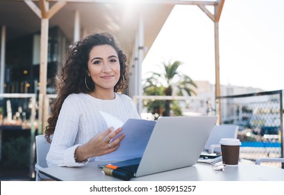Portrait of skilled female student with paper documents sitting at table with modern laptop computer and posing, successful freelancer with financial report looking at camera during distance job