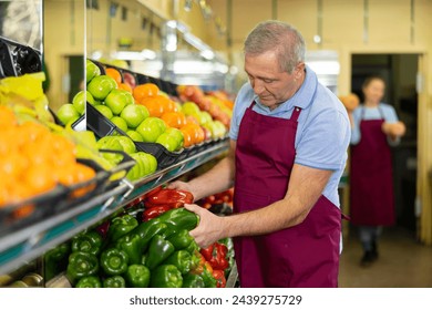 Portrait of skilled mature salesman in apron checking bell peppers before selling in grocery supermarket - Powered by Shutterstock