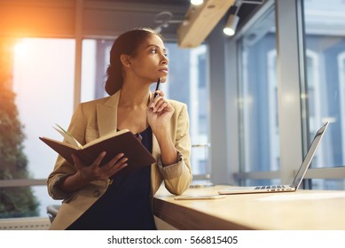 Portrait of skillful afro american female executive thinking over working schedule for employee writing report into notebook while using laptop computer and wireless connection in coworking space  - Shutterstock ID 566815405