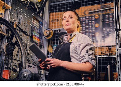 Portrait Of Skillful Old Woman Mechanic Holding Tablet Around Bench And Bicycle In Workshop.