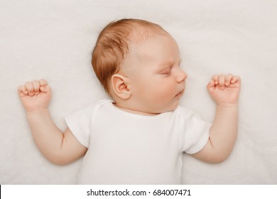 Portrait of sleeping cute Caucasian little baby newborn in white clothes lying on bed with his hands up. Lifestyle candid real concept.