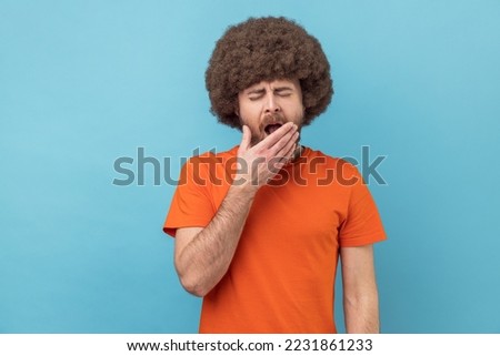 Portrait of sleepy man with Afro hairstyle wearing orange T-shirt yawning and covering mouth with hand, feeling exhausted, lack of sleep. Indoor studio shot isolated on blue background.
