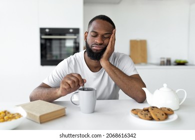 Portrait of sleepy young African American man sitting at dining table in kitchen, stirring coffee with closed eyes, having breakfast selective focus. Early Wake Up, Insomnia, Lack Of Sleep