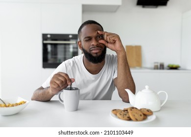 Portrait of sleepy young African American man trying to open his eye with fingers, sitting at dining table in kitchen, stirring coffee. Early Wake Up, Insomnia, Lack Of Sleep, Morning Fatigue - Powered by Shutterstock