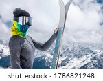 Portrait of a slender girl in a buff and a balaclava in a ski mask and a hat with a closed face next to skis against the backdrop of snow-covered epic mountains.