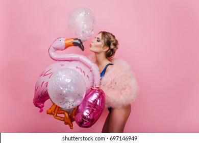 Portrait of slim graceful girl with short blonde hair kissing pink inflatable flamingo. Gorgeous cheerful young woman in fluffy coat having fun with sparkle balloons, waiting for party