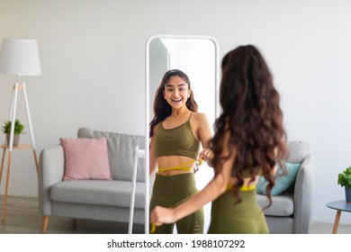 Portrait of slim Indian woman measuring her waist in front of mirror at home, free space. Beautiful Eastern lady overjoyed with result of her weight loss diet. Healthy eating concept
