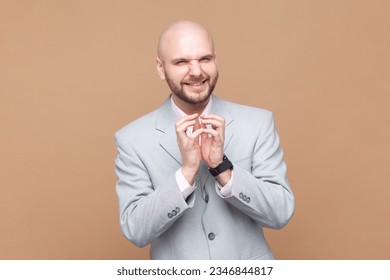 Portrait of sly smiling bald bearded man looking at camera with cunning facial expression, planning devil prank, wearing gray jacket. Indoor studio shot isolated on brown background.