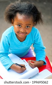 Portrait Of Small African Kid Writing In Notebook At Desk.