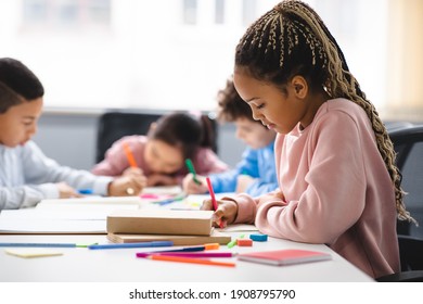 Portrait of small black girl with afro braids sitting at table in classroom at primary school or kindergarten, writing or drawing in notebook. Reopening and return back to school after lockdown - Shutterstock ID 1908795790