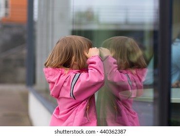 Portrait Of Small Blond Girl In Preschool Age Looking Through Transparent Glass Window To Shop
