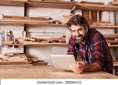 Portrait of a small business owner smiling at the camera, resting on his workbench and holding a digital tablet, with shelves of wood behind him in his woodwork studio - Powered by Shutterstock