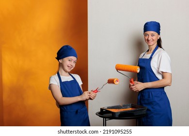 Portrait Of Small Family Painting Room Walls With Orange Paint And Roller Paintbrush, Working On Apartment Redecoration. Woman With Little Girl Using Dye Color To Renovate House.