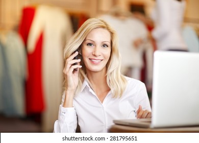 Portrait Of Small Fashion Store Owner. Busy Businesswoman Sitting In Her Designer Shop And Making Call While And Using Laptop.