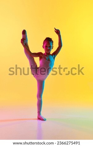 Portrait of small flexible child ballerina dancer girl in sport swimsuit and in pointe white legging posing holding one leg and hand in mixed neon light in choreography hall. Children ballet concept