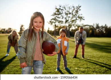 Portrait Of Small Girl Holding An Oval Brown Leather Rugby Ball While Playing American Football With Her Parents And Brother In Park. Family And Kids, Nature Concept. Horizontal Shot