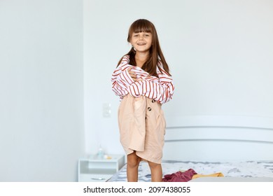 Portrait Of Small Girl Wearing Large Striped Shirt And Beige Short Pretending To Be A Grown Up Woman. Girl Child Posing In Oversized Clothes Indoor While Standing On Bed.