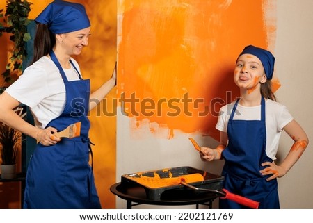 Portrait of small kid with tongue out fooling around and painting walls with orange color paint to do housework renovation wiht mother. Little girl playing with paint, being messy.
