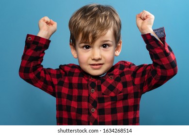 Portrait Of Small Little Caucasian Boy Young Child Kid Standing In Front Of The Blue Wall Background Wearing Red And Black Shirt Looking To Camera In Studio Holding Fists In Flexing Muscles Victory