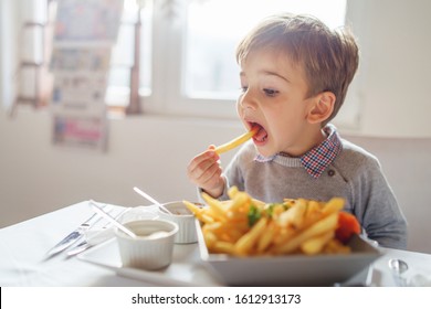 Portrait Of Small Little Cute Caucasian Boy Kid Eating French Fries Potato Chips At The Table In The Restaurant Or At Home Three Or Four Years Old