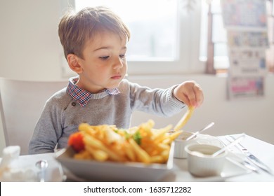 Portrait Of Small Little Cute Caucasian Boy Kid Eating French Fries Potato Chips At The Table In The Restaurant Or At Home Three Or Four Years Old