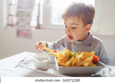 Portrait Of Small Little Cute Caucasian Boy Kid Eating French Fries Potato Chips At The Table In The Restaurant Or At Home Three Or Four Years Old