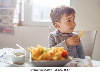 Portrait Of Small Little Cute Caucasian Boy Kid Eating French Fries Potato Chips At The Table In The Restaurant Or At Home Three Or Four Years Old