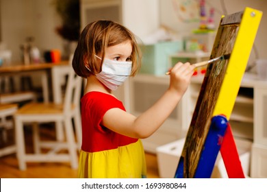 Portrait Of A Small Preschool Child With Face Mask Against Coronavirus, Painting At An Art Easel At Home