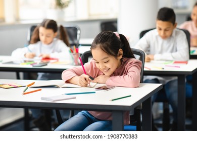 Portrait of small smiling asian girl sitting at table in classroom at primary school or kindergarten, writing or drawing in notebook. Reopening and return back to school after coronavirus quarantine - Powered by Shutterstock