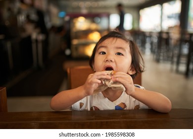 Portrait Of Small Two Years Old Boy Child Sitting In The Chair Enjoying A Piece Of Delicious Donut. Facial Expression Is Showing Very Appetizing And Greedy. Shot With Short Depth Of Field In Focus.