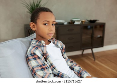 Portrait of smart charismatic and serious African American boy of 7-9 years sitting in living room on grey couch, against stylish interior, looking at camera with big beautiful brown eyes