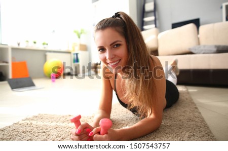 Portrait of smart female yoga trainer looking at camera with gladness and calmness and holding pink dumbbells in tender hands. Gorgeous girl lying on floor. Training at home concept