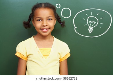 Portrait Of Smart Girl By The Blackboard Thinking Of Light Idea And Looking At Camera