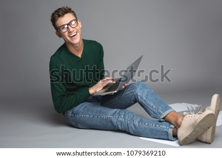 Portrait of smart guy sitting with computer on knees and rejoicing. He is wearing pair of spectacles