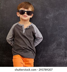 Portrait Of Smart Little Boy Wearing Sunglasses Smirking. Cool Kid In Shades Leaning On A Blackboard Looking At Camera Smiling With Copy Space.