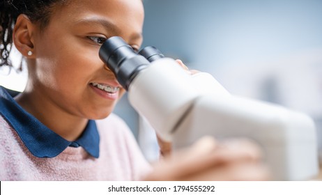 Portrait Of Smart Little Schoolgirl Looking Under The Microscope. In Elementary School Classroom Cute Girl Uses Microscope. STEM (science, Technology, Engineering And Mathematics) Education Program