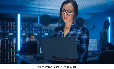 Portrait of a Smart Seductive Young Woman Wearing Glasses Holds Laptop. In the Background Technical Department Office with Specialists Working and Functional Data Server Racks