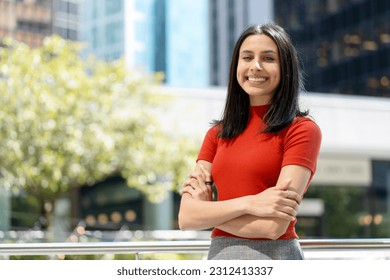 Portrait of smart smiling Indian student with arms crossed looking at camera in university campus, education concept. Beautiful confident asian woman wearing red stylish t shirt standing on street 