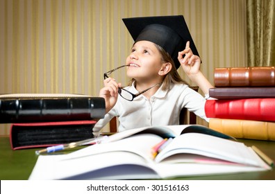 Portrait of smart thoughtful girl posing at desk in graduation cap