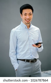 Portrait Of A Smartly Dressed Chinese Asian Man Holding His Smartphone And Smiling At The Camera. He Is Standing Against A Plain Grey Blue Background And Is Relaxed, Natural And Authentic. 