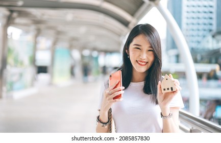 Portrait Of Smile Young Adult Business Working Asian Woman. Hand Holding Mobile Phone And House Model For Online Insurance Application Concept. Background With Copy Space On Day.