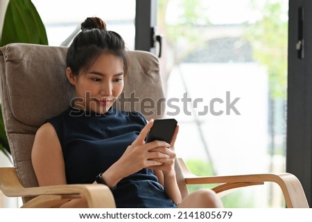 A portrait of a smiley Asian woman sitting on a chair in a living room  using a smart phone, for lifestyle, home and technology concept.