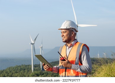 Portrait of smiley handsome asian man engineer wearing safety helmet, reflective vest holding tablet and walkie talkie with wind turbine propeller background. Environmental friendly concept.