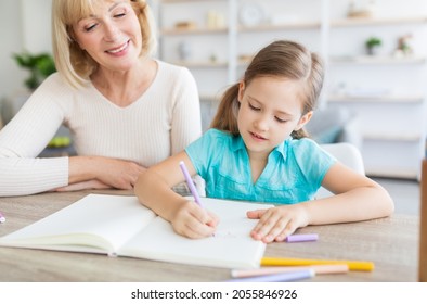 Portrait of smilig mature woman helping little girl with homework, spending free time with small preshool grandchild, sitting together at table, drawing pictures using colorful pencils and markers