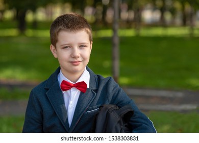 Portrait Of A Smiling 10 Year Old Boy In A School Jacket With A Red Bow Tie And Backpack. Outdoors In The Park.